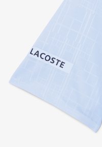 Ljusblå handduk med mönstrad struktur och ett vitt band med svart text som säger "LACOSTE" tydligt synlig.