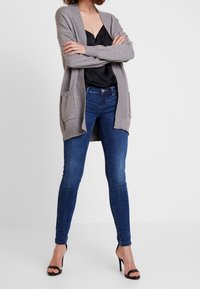 Grauer Strickcardigan mit offenem Vorderteil, zwei Taschen, getragen über einem schwarzen Satinoberteil und blauen engen Jeans, kombiniert mit schwarzen Absatzsandalen.