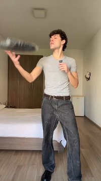 Young man in gray shirt and dark jeans tossing a black plastic-wrapped item in a bright bedroom with bed and wardrobe visible.