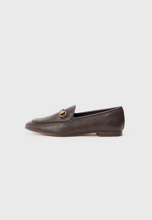 Mocassins en cuir marron avec un bout pointu, ornés d'une accent en doré à l'avant et d'une semelle plate. Texture lisse dans l'ensemble.