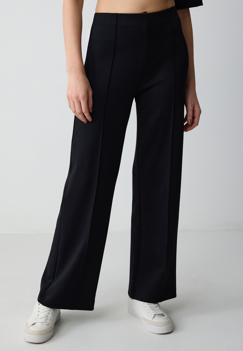 Zwarte wijde broek van gladde stof, met een hoge taille en plooien aan de voorkant. Gecombineerd met witte sneakers voor een casual uitstraling.