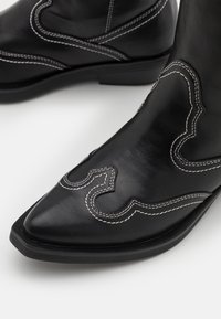 Zwarte leren laarzen met een puntige neus, voorzien van witte stiksels in een gebogen patroon langs de bovenkant en een lage, platte zool.