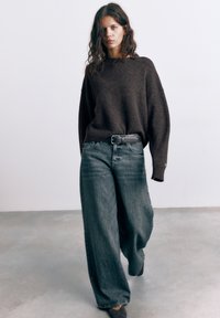 Donna con capelli lunghi e ondulati che indossa un maglione scuro oversize, jeans larghi sbiaditi e una cintura nera, che cammina su un pavimento di cemento grigio.