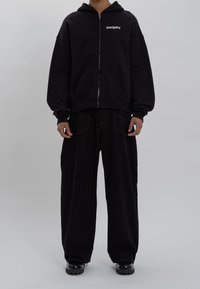 Felpa nera con zip in un tessuto morbido, dotata di tasca frontale e logo. Abbinata a pantaloni neri larghi e scarpe nere chunky.