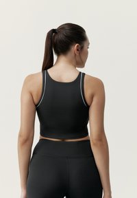 Zwart atletisch crop top met brede bandjes en een aangesloten ontwerp. Textuurstof met contrasterende stiksels. Draagt in combinatie met bijpassende zwarte leggings.