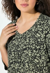 Schwarze Bluse mit Blumenmuster, V-Ausschnitt, kurzen Ärmeln und grünem Blumenprint auf weicher Stoffqualität, mit sichtbaren Faltendetails.
