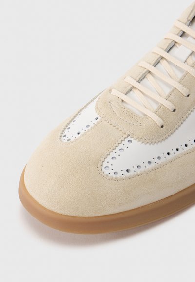 Μπεζ σουέτ αθλητικό παπούτσι με λευκές δερμάτινες λεπτομέρειες, διακοσμητικό διάτρητο σχέδιο και σόλα από καουτσούκ τύπου γκαμ. Διαθέτει τυπικά κορδόνια και στρ ογγυλεμένη μύτη.