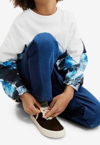 Sudadera blanca con mangas abstractas en azul y negro, combinada con pantalones de mezclilla azules. Zapatillas marrones con suela blanca, con los cordones atados.