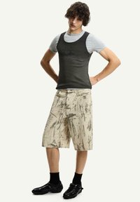 Grå ärmlös tanktopp under en svart skjorta, beige shorts med mörka abstrakta mönster, svarta skor och ankelsockar.