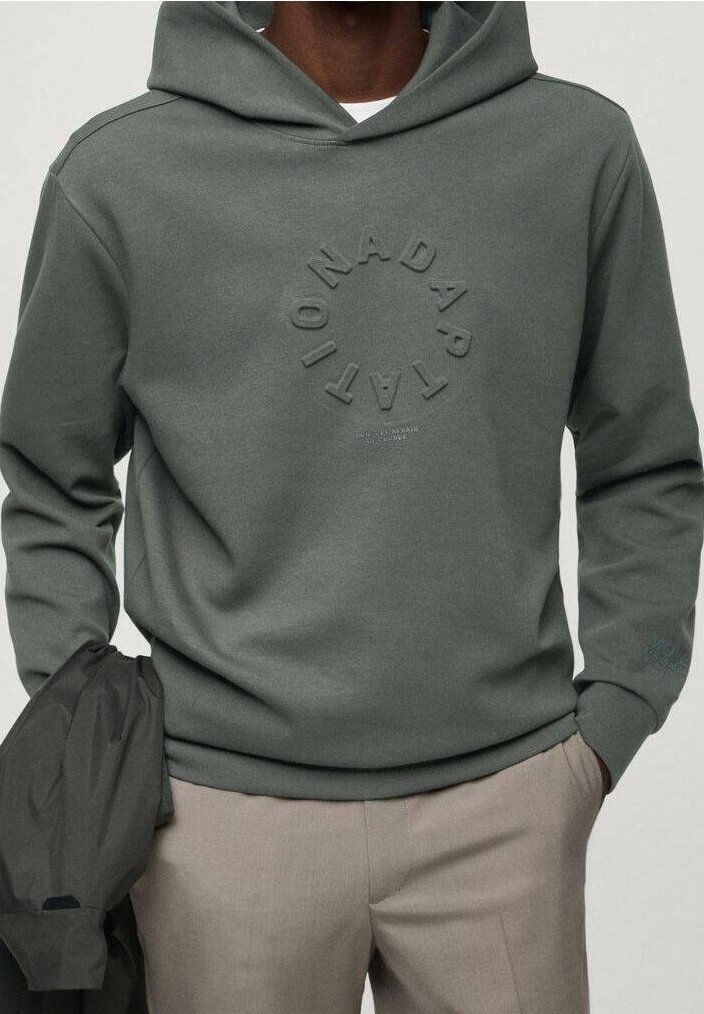 Homme portant un sweat à capuche vert pâle avec un texte circulaire en relief et un pantalon beige, tenant une veste sombre dans une main.