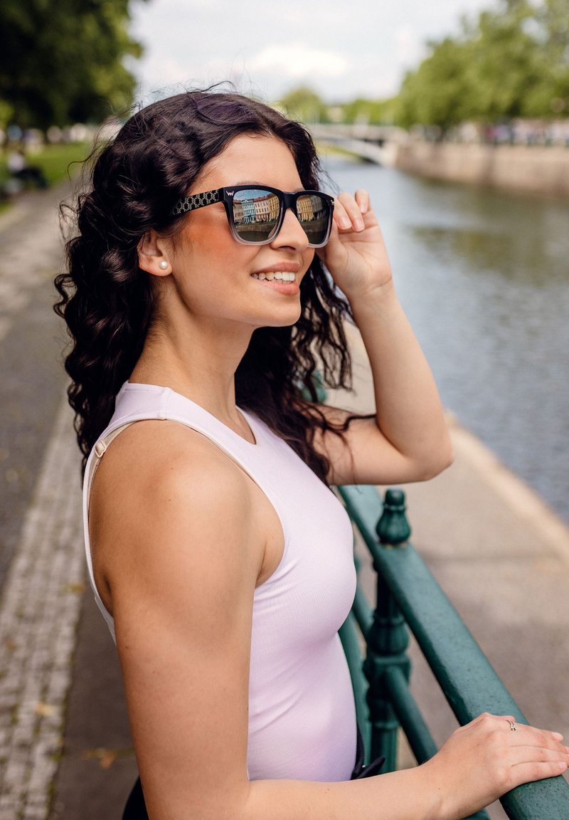 Černé sluneční brýle s geometrickým vzorem na stranách, které nosí žena s kudrnatými vlasy. Stojí u zeleného zábradlí poblíž vodní plochy.