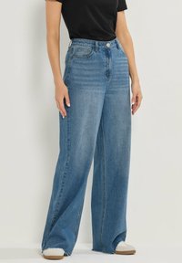 Jean large bleu clair en denim. Taille haute avec poches avant et ourlet effiloché. Associé à des baskets blanches.