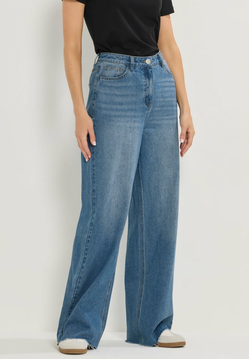 Jean large bleu clair en denim. Taille haute avec poches avant et ourlet effiloché. Associé à des baskets blanches.