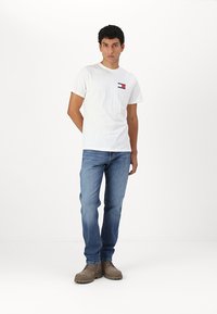 White cotton t-shirt with a small red and blue logo on the left chest, paired with light blue denim jeans and brown boots.