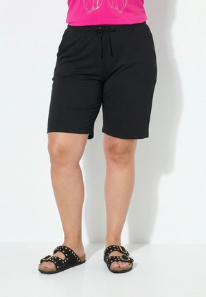 Person iført sorte shorts med snøre og sorte nitte-sandaler med spænder, stående mod en ensfarvet hvid baggrund.