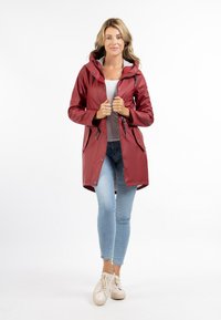 Water-resistant red raincoat with a hood and snap buttons, featuring side pockets and a cinched waist, worn over blue jeans and beige shoes.