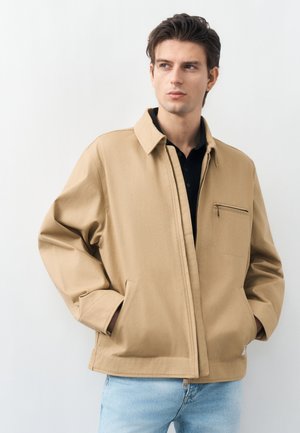 Młody mężczyzna w beżowej kurtce na zamek, z rękami w kieszeniach, patrzący w bok, stojący na tle jednolitego jasnego tła.