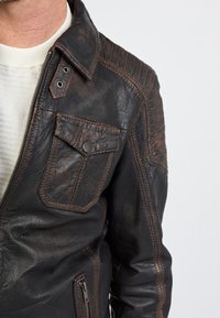 Braune Lederjacke mit strukturiertem Finish, sichtbaren Nähten, seitlicher Reißverschlusstasche und Brusttasche mit Druckknopfverschluss. Gerippte Schulterdetails.