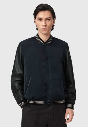 Młody mężczyzna zwrócony przodem, ubrany w czarną kurtkę varsity z skórzanymi rękawami oraz prążkowanym kołnierzem, mankietami i pasem na biodrach.