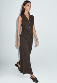 Femme portant un gilet sans manches marron foncé boutonné avec un pantalon large assorti et des chaussures sombres à enfiler, marchant devant un fond gris clair.