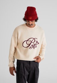 Bărbat îmbrăcat cu un pulover crem cu un design floral maroon în formă de "P", pantaloni închiși plisati și o căciulă tricotată roșie, privindu-se în jos cu o mână în buzunar.