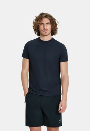 Uomo con capelli ricci che indossa una maglietta a maniche corte blu scuro tinta unita e pantaloncini abbinati, in piedi con una mano in tasca.