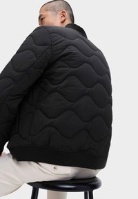 Svart kviltad bomberjacka med en vågmönster, ribbad krage och mudd. Modellen sitter på en svart pall och visar jackans textur.