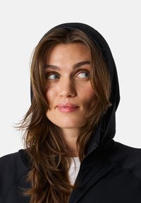 Svart zip-up hoodie med huva, tillverkad av ett slätt material. Har dragkedjestängning, tight passform och bärs över en vit skjorta.