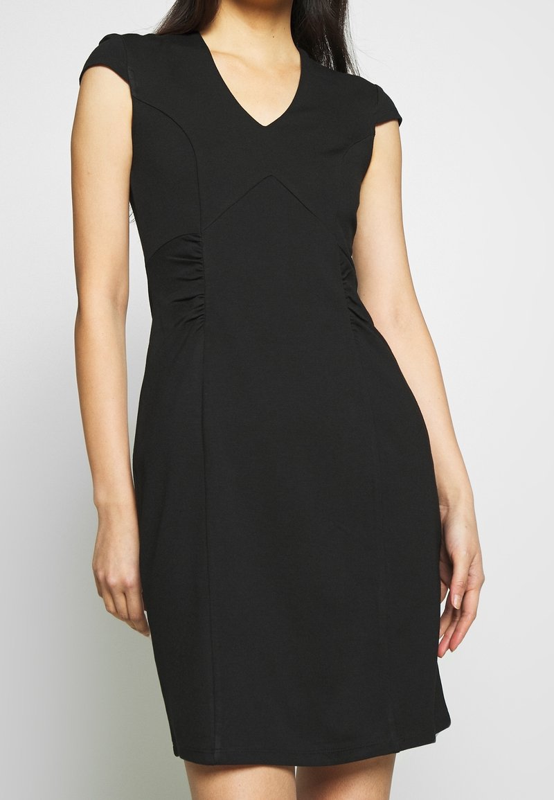 Femme portant une robe noire ajustée à manches courtes et un décolleté en V, se tenant devant un fond clair uni.
