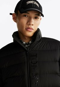 Czarna kurtka puchowa z wysokim kołnierzem, o błyszczącej fakturze z subtelnym wytłoczonym logo "TOMMY"; noszona z czapką w kolorze czarnym z napisem "TOMMY JEANS".