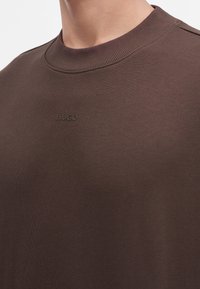 Gros plan d'une personne portant un sweat-shirt marron à col rond avec un petit texte en relief "HUGO" sur la poitrine.