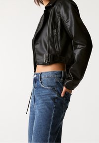 Chaqueta corta de cuero negro con cuello alto, cremalleras metálicas y hebilla de cinturón. Combinada con jeans de mezclilla azul con efecto desgastado.