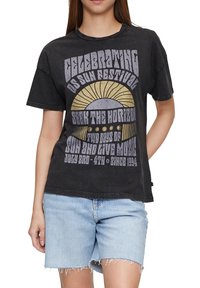 Žena v černém festivalovém grafickém tričku se sluncem a textem, doplněném o světle modré roztrhané džíny.