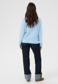 Hosszú barna hajú nő, világoskék pulóverben, sötét, felhajtott farmernadrágban és barna cipőben áll, egyszínű háttér előtt.