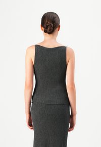 Kvinne med mørkt hår i en ryddig knute, iført en ermeløs, teksturert svart topp og matchende skjørt, vendt bort fra en enkel hvit bakgrunn.