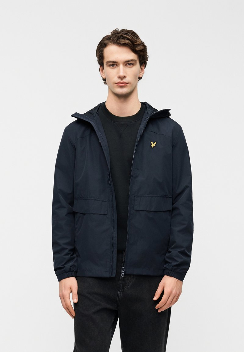 Ναυτικό ελαφρύ μπουφάν με κουκούλα, δύο εμπρόσθιες τσέπες και ελαστικά μανσέτα· διαθέτει μικρό χρυσό λογότυπο στο αριστερό στήθος.