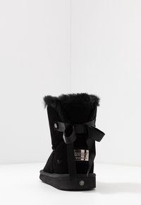 Botín negro de ante hasta el tobillo con un cuello forrado de piel, presenta un lazo decorativo de cinta negra y una suela de goma. Etiqueta de tamaño visible en el costado.
