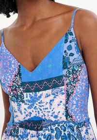 Femme portant une robe sans manches avec des motifs floraux et géométriques en patchwork bleu, rose et vert, ainsi que des bretelles fines.