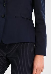 Granatowa marynarka wykonana z gładkiej tkaniny, z pojedynczą kieszenią na piersi, dopasowanym krojem i zapięciami na guziki. Połączona z spodniami w prążki.