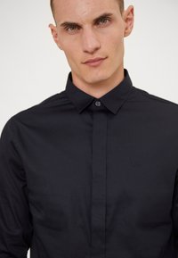 Camisa de botones azul marino oscuro hecha de tela suave, con cuello, manga larga y un sutil logo grabado en el pecho.