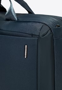 Donkerblauwe stoffen tas met een gladde textuur, stevige hengsels en een zilveren merketiket. Rits sluiting en versterkte naden zijn zichtbaar.