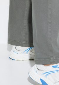 Un pantalon large gris clair tombe au-dessus de chaussures de sport blanches et bleues avec des détails en mesh et des rayures contrastées. Hems cousus visibles.