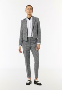Maßgeschneiderter grauer und weißer gepunkteter Anzug mit einem taillierten Blazer, engen Hosen und einem weißen Hemd darunter. Schwarz-weiße Loafers runden den Look ab.