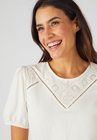Blusa blanca de manga corta con un yugo de encaje bordado detallado y recortes, creando un patrón floral alrededor del escote.
