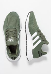 Gröna nätliknande sportskor med vitt tre-stripes-mönster, texturerad överdel, vadderad tunga och kontrasterande svart sula.