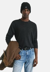 Zwarte gebreide muts, donkergrijs langebloes en blauwe denim jeans. Bruine jas over één schouder, wat een casual look laat zien.
