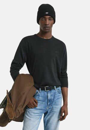Schwarze Strickmütze, dunkelgraues Langarmshirt und blaue Jeans. Braune Jacke lässig über eine Schulter geworfen, was einen entspannten Look zeigt.