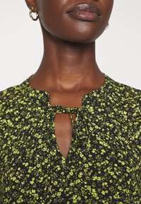Blouse noire avec un motif floral en vert néon, dotée d'un détail de cou en clé avec un nœud. Le tissu semble léger avec une texture douce.