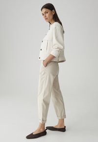 Femme en pantalon beige, veste crème boutonnée avec poches et ballerines marron foncé, debout de profil avec les mains dans les poches sur un fond gris uni.