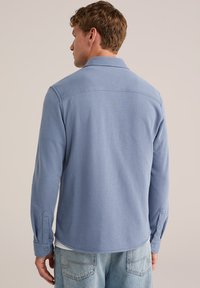 Camisa azul de manga larga hecha de tejido suave, con cuello, puños abotonados y dobladillo curvado. Vista trasera con detalles de costura mínimos.